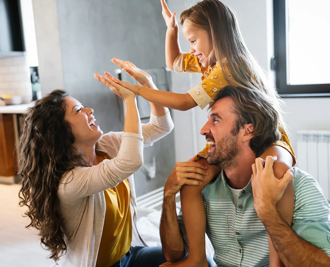 Caucasian family sitting on the floor where the father is holding their daughter on his shoulders why she gives high-fives to the mother. This image depicts a young family sitting comfortably in their own home.