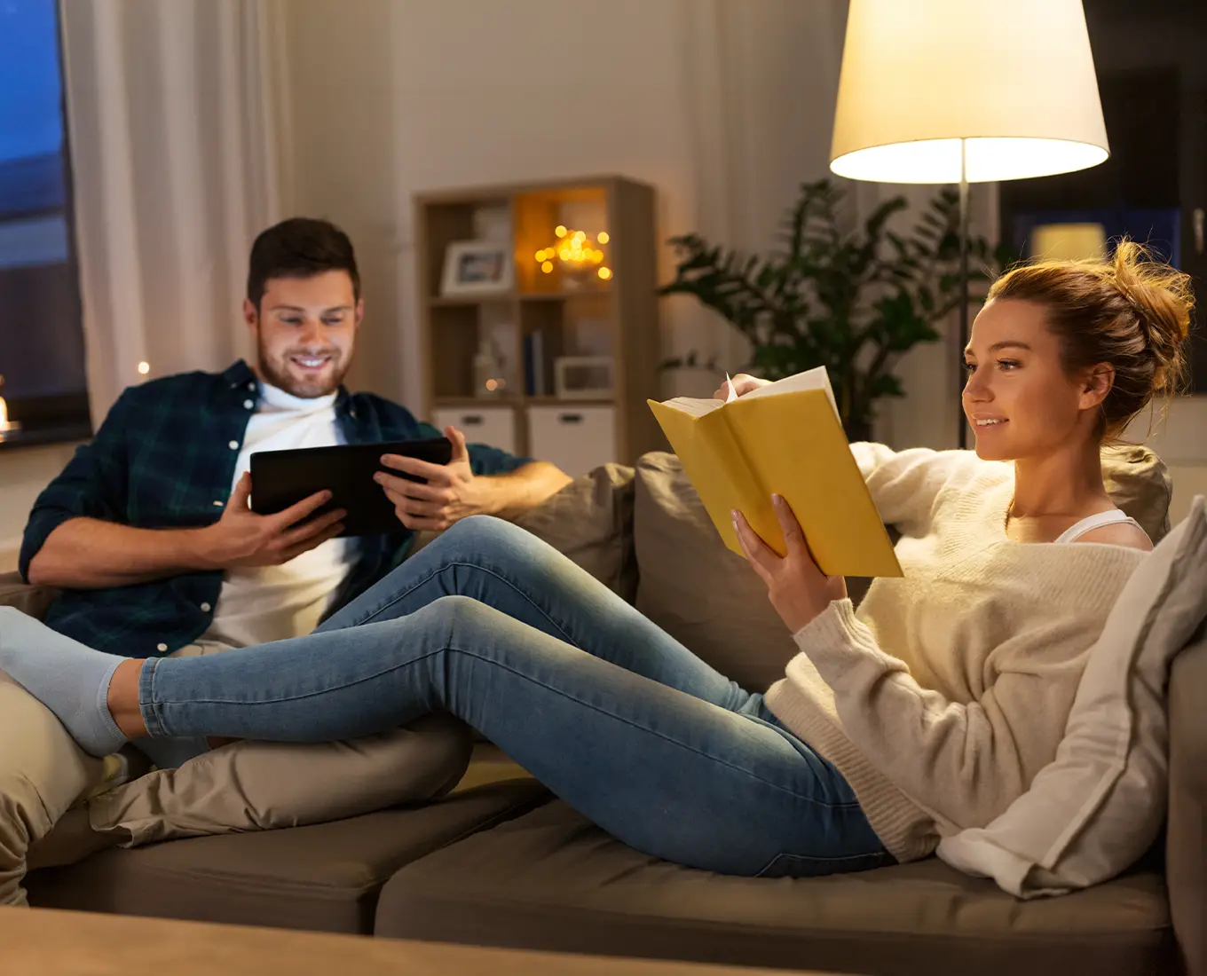Couple relaxing on a couch at night with lights on, the woman reading a book and the man using a tablet, illustrating reliable generator power keeping the home running during a power outage.