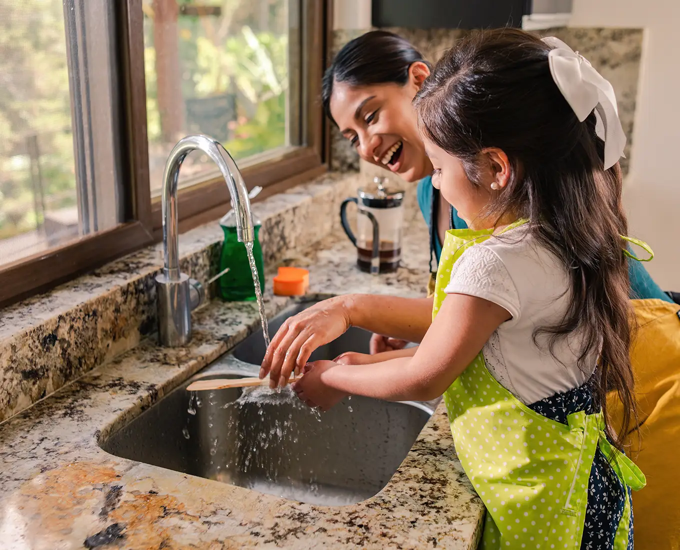 Mother and daughter smiling while washing dishes together at a kitchen sink with running hot water, illustrating reliable hot water from a home water heater for everyday use.